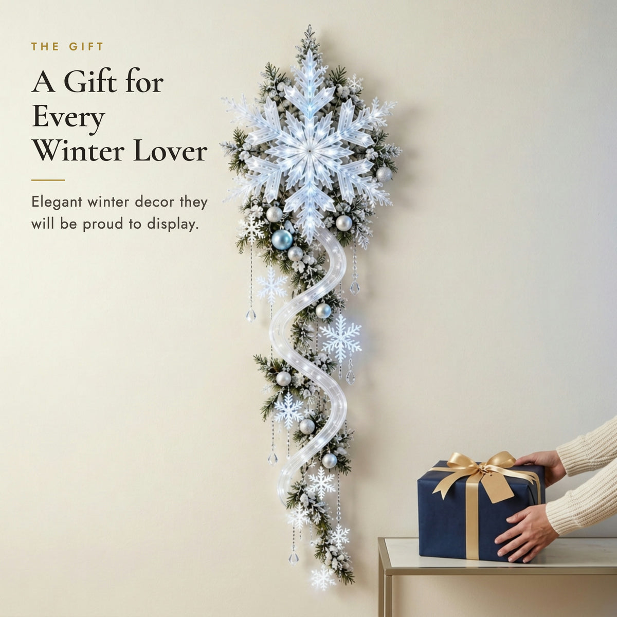 Hands placing a navy gift box with gold ribbon below the lit snowflake wall decor